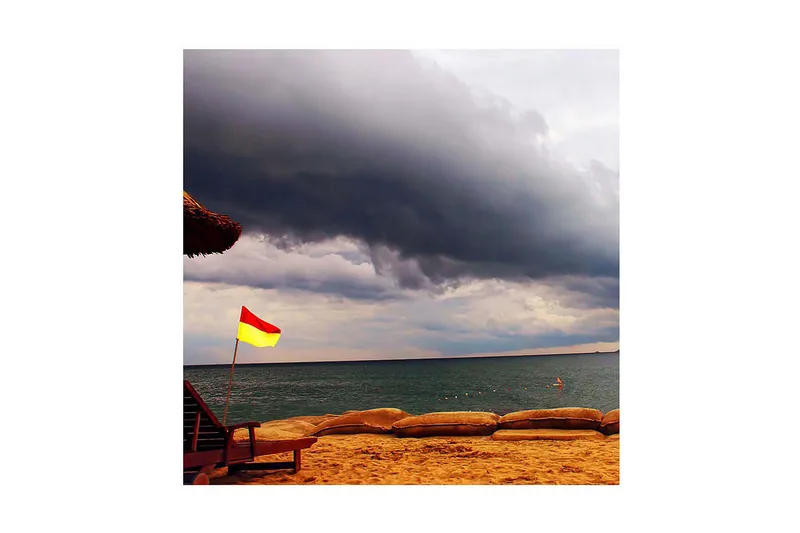 Canvas-taulu 45x45 cm - Punainen ja keltainen lippu rannalla, taustalla tummia pilviä, Tummanharmaa / Punainen / Keltainen