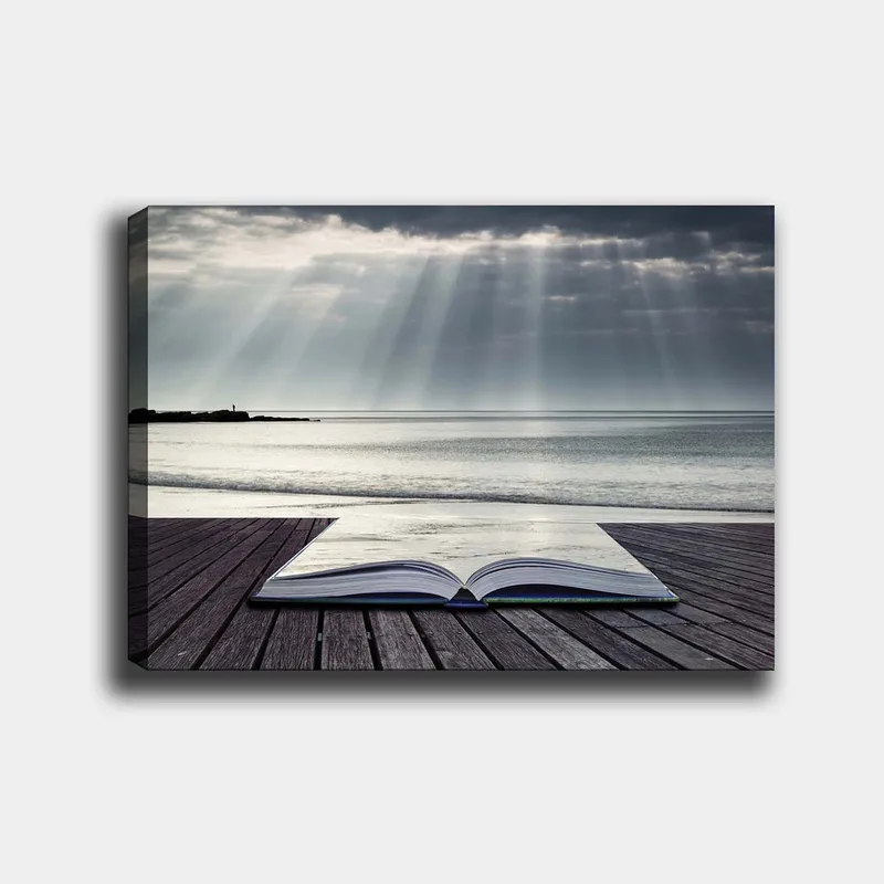 Canvas taulu 70x100 cm - Avoin kirja puisella laiturilla meren rannalla, pilvisen taivaan pehmeiden valonsäteiden ympäröimänä, Harmaa / Sininen / Valkoinen