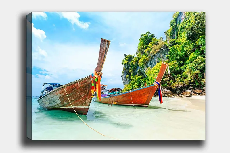 Canvas taulu 70x100 cm - Kaksi perinteistä thaimaalaista pitkävenettä rannalla, taustalla kiviä ja rehevää kasvillisuutta, Ruskea / Vihreä / Sininen