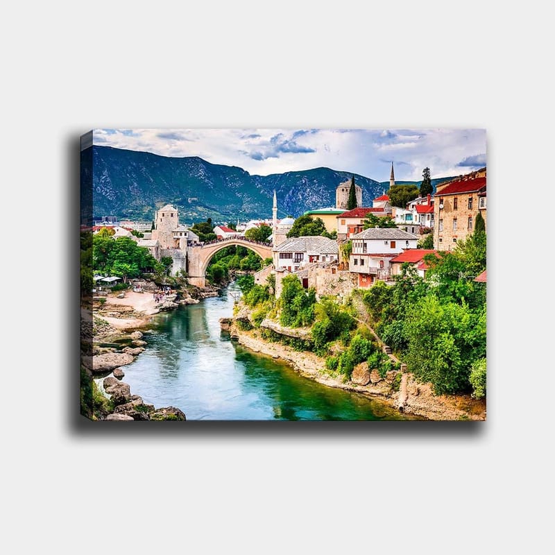 Canvas taulu 70x100 cm - Kaunis näkymä historiallisesta joen ylittävästä sillasta, jota ympäröivät viehättävät talot ja rehevä vehreys., Vihreä / Sininen / Punainen