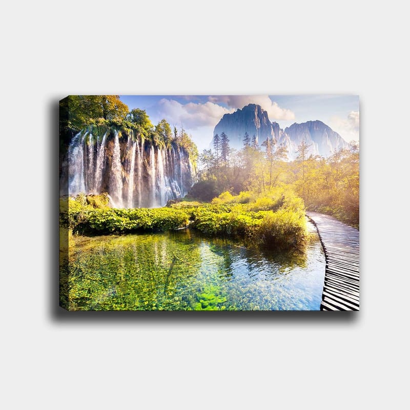 Canvas taulu 70x100 cm - Upea vesiputousmaisema rehevän kasvillisuuden ja majesteettisten vuorten ympäröimänä, Vihreä / Sininen / Keltainen