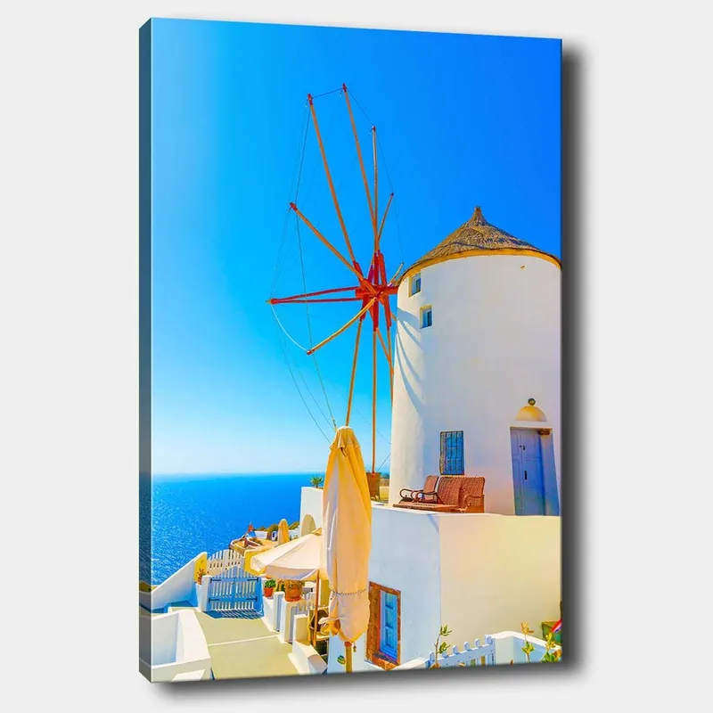 Canvas taulu 70x100 cm - Valkoinen tuulimylly punaisella roottorilla kalliolla meren rannalla, kirkkaan sinisen taivaan ja meren ympäröimänä, Valkoinen / Punainen / Sininen
