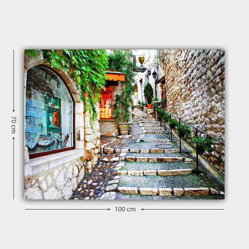 Canvas taulu 70x100 cm - Viehättävä kuja, jonka varrella on kiviä ja vehreyttä, ja jota ympäröivät historialliset rakennukset. - Vihreä / Beige / Oranssi - Sisustustuotteet - Taulu & taide - Canvas-taulu