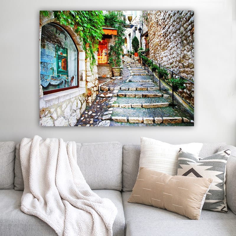 Canvas taulu 70x100 cm - Viehättävä kuja, jonka varrella on kiviä ja vehreyttä, ja jota ympäröivät historialliset rakennukset. - Vihreä / Beige / Oranssi - Sisustustuotteet - Taulu & taide - Canvas-taulu