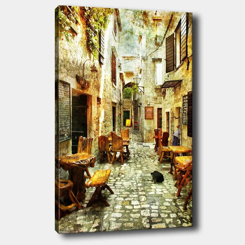 Canvas taulu 70x100 cm - Viehättävä kuja, jossa on puisia pöytiä ja tuoleja, vanhojen kivitalojen ympäröimänä, Ruskea / Harmaa / Vihreä