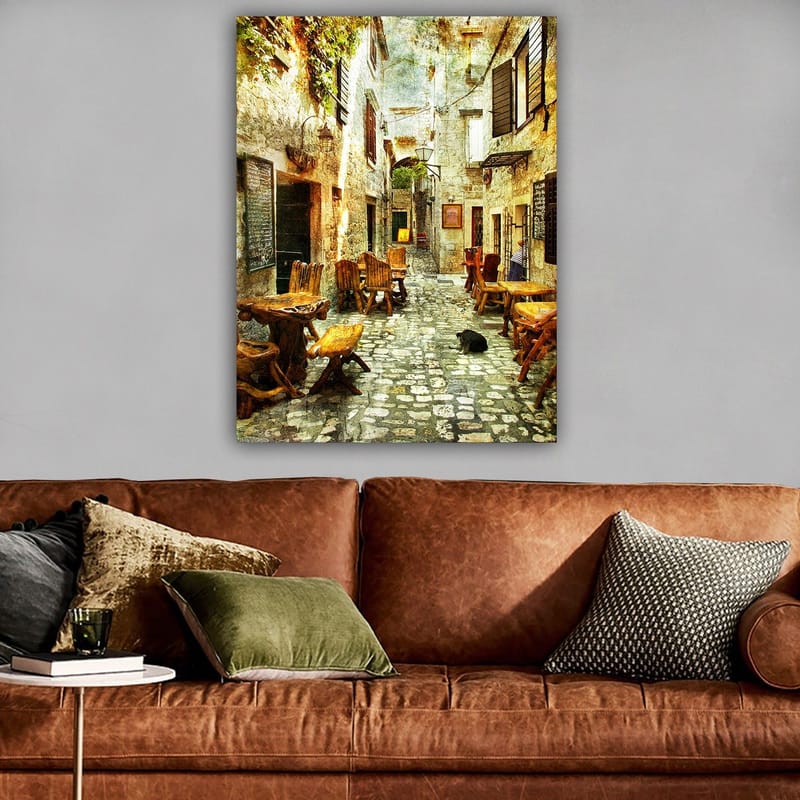 Canvas taulu 70x100 cm - Viehättävä kuja, jossa on puisia pöytiä ja tuoleja, vanhojen kivitalojen ympäröimänä - Ruskea / Harmaa / Vihreä - Sisustustuotteet - Taulu & taide - Canvas-taulu