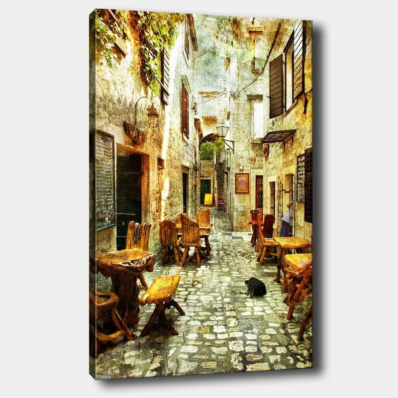 Canvas taulu 70x100 cm - Viehättävä kuja, jossa on puisia pöytiä ja tuoleja, vanhojen kivitalojen ympäröimänä, Ruskea / Harmaa / Vihreä