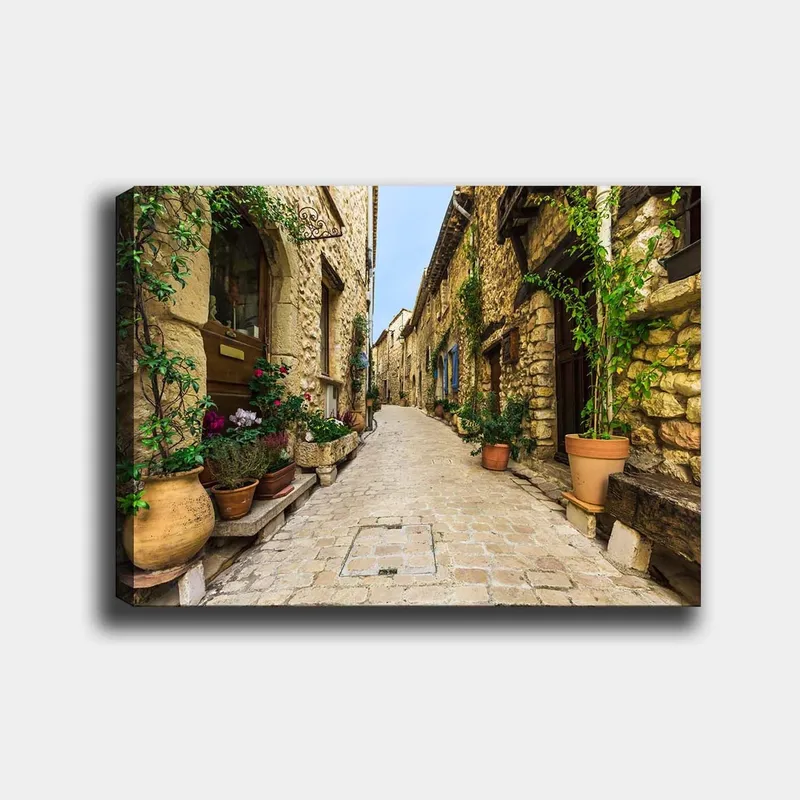 Canvas taulu 70x100 cm - Viehättävä kuja vanhassa kylässä kivitalojen ja kukkaruukkujen ympäröimänä, Beige / Vihreä / Terrakotta