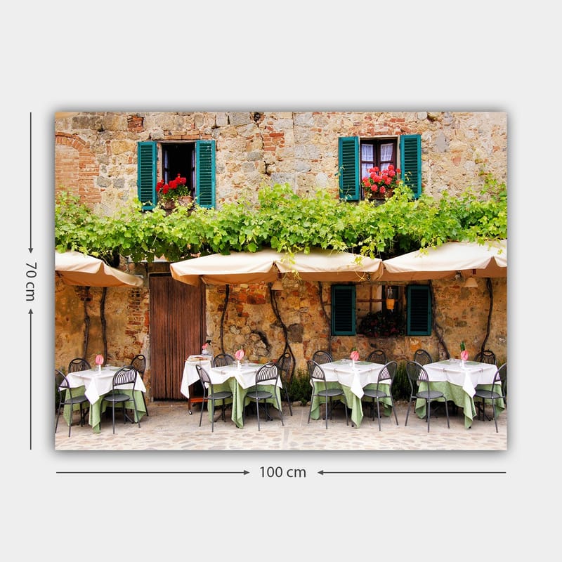 Canvas taulu 70x100 cm - Viehättävä ulkoruokailutila valkoisine pöydineen ja vihreine pöytäliinoineen vanhan kivitalon edessä, jossa on tummanvihreät ikkunaluukut - Tummanvihreä / Valkoinen / Tiilenpunainen - Sisustustuotteet - Taulu & taide - Canvas-taulu
