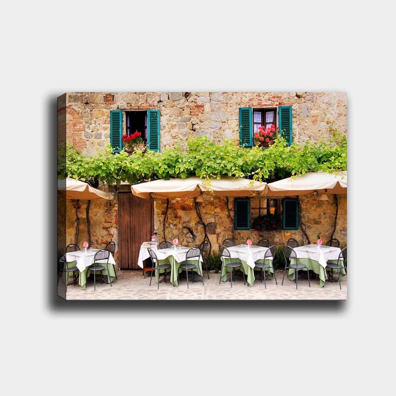 Canvas taulu 70x100 cm - Viehättävä ulkoruokailutila valkoisine pöydineen ja vihreine pöytäliinoineen vanhan kivitalon edessä, jossa on tummanvihreät ikkunaluukut, Tummanvihreä / Valkoinen / Tiilenpunainen