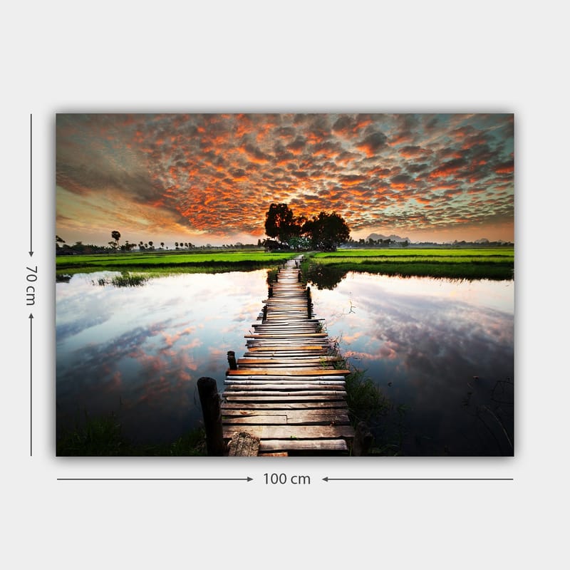 Canvas taulu 70x100 cm - Puinen silta, joka ylittää peilimäisen veden pinnan, rehevän vehreyden ja dramaattisen auringonlaskun ympäröimänä - Oranssi / Vihreä / Sininen - Sisustustuotteet - Taulu & taide - Canvas-taulu