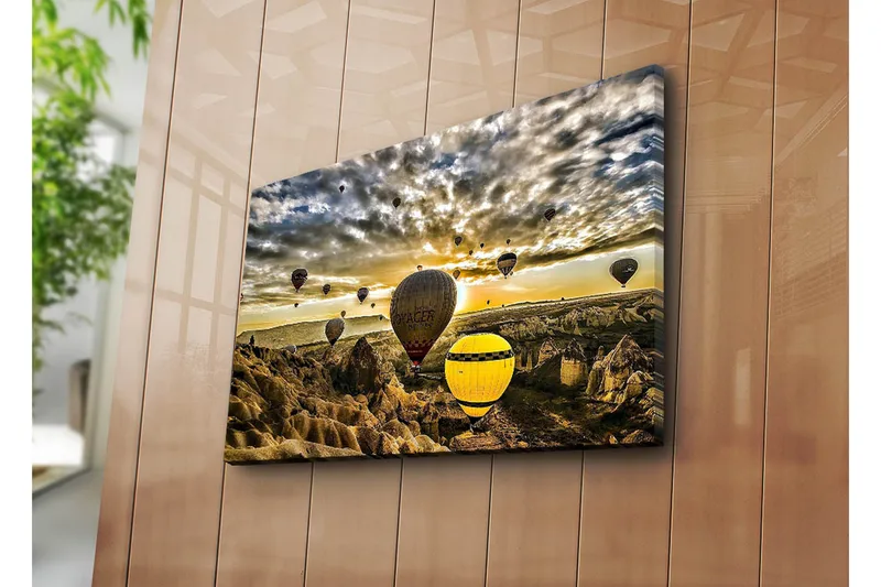 Canvas-taulu 70x45 cm - Useita värikkäitä kuumailmapalloja leijuu dramaattisen maiseman yllä auringonlaskun aikaan, Keltainen / Harmaa / Ruskea
